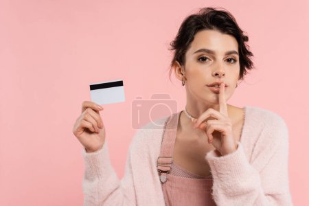 joven morena mujer en acogedor cárdigan mostrando signo de silencio mientras sostiene la tarjeta de crédito aislado en rosa