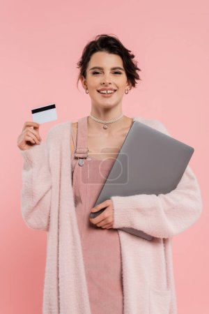 Foto de Mujer morena feliz con ordenador portátil y tarjeta de crédito mirando a la cámara aislada en rosa - Imagen libre de derechos