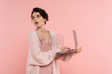 astonished brunette woman with laptop looking back isolated on pink