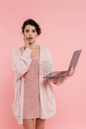 worried woman with laptop touching face and looking at camera isolated on pink