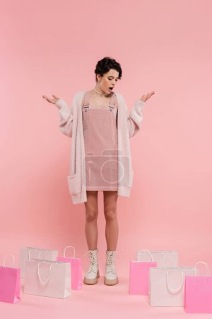 full length of amazed woman showing wow gesture while looking at shopping bags on pink background