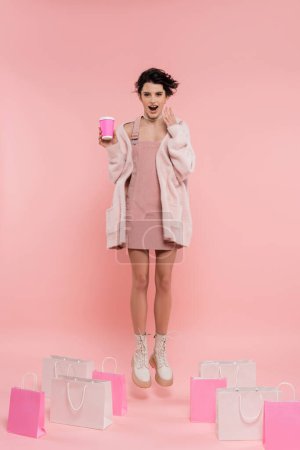 longitud completa de la mujer asombrada con café para ir levitando cerca de bolsas de compras sobre fondo rosa