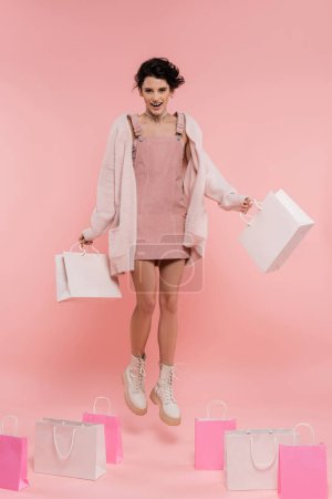 volle Länge der fröhlichen Frau in Trägerkleid und flauschige Strickjacke schwebt mit Einkaufstaschen auf rosa Hintergrund