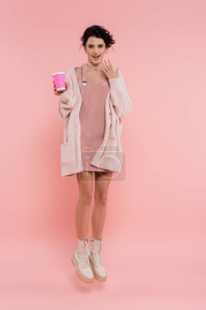 full length of excited woman in strap dress and cozy cardigan levitating with paper cup on pink background