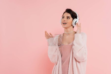 Téléchargez les photos : Femme étonnante et joyeuse dans les écouteurs sans fil regardant loin isolé sur rose - en image libre de droit