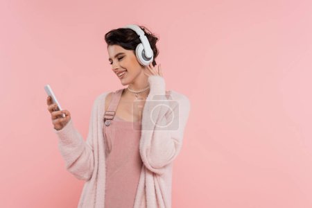 happy brunette woman in wireless headphones looking at mobile phone isolated on pink