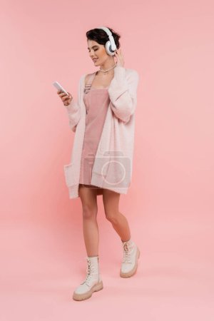 full length of fashionable woman in wireless headphones walking with smartphone on pink 