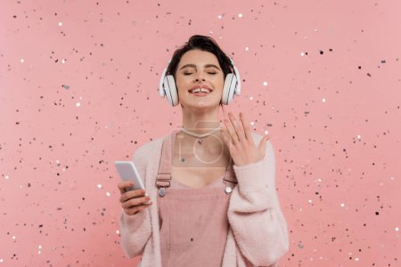 mujer alegre con los ojos cerrados sosteniendo el teléfono celular y escuchando música en auriculares inalámbricos cerca de confeti en rosa 