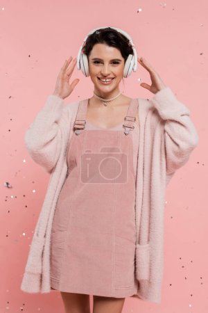 Téléchargez les photos : Femme heureuse en robe de sangle et cardigan chaud tenant les mains près des écouteurs sans fil sur fond rose - en image libre de droit