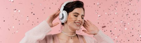 Lächelnde brünette Frau mit geschlossenen Augen, die Musik in drahtlosen Kopfhörern in der Nähe von Konfetti auf rosa Hintergrund hört, Banner