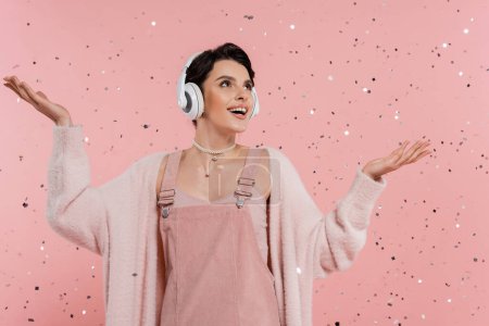 Téléchargez les photos : Femme étonnante et heureuse dans les écouteurs sans fil debout sous confettis sur fond rose - en image libre de droit