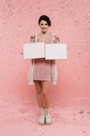 volle Länge der glücklichen und trendigen Frau mit Einkaufstaschen, die unter Konfetti auf rosa Hintergrund stehen