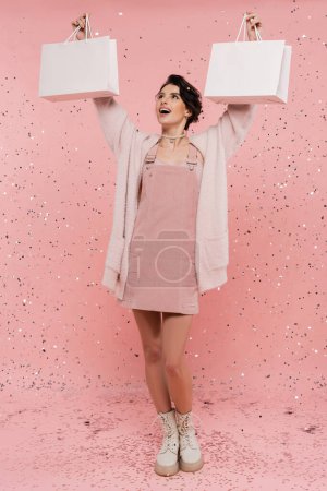 Photo for Full length of excited and fashionable woman holding shopping bags in raised hands under festive confetti on pink background - Royalty Free Image