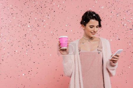 Téléchargez les photos : Femme brune branchée avec café pour aller messagerie sur smartphone sous confettis sur fond rose - en image libre de droit