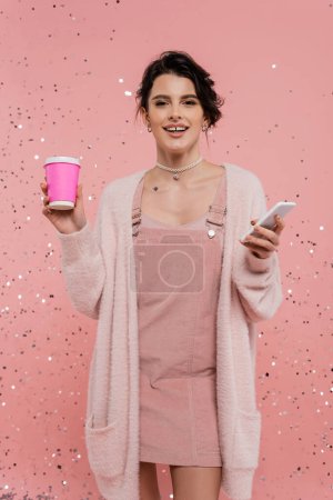 Téléchargez les photos : Femme brune heureuse avec tasse en papier et téléphone mobile en regardant la caméra sur fond rose - en image libre de droit