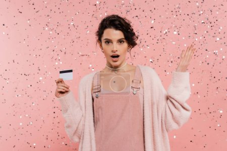 Téléchargez les photos : Femme étonnée en robe de sangle et cardigan tenant carte de crédit et pointant avec la main sous confettis sur fond rose - en image libre de droit