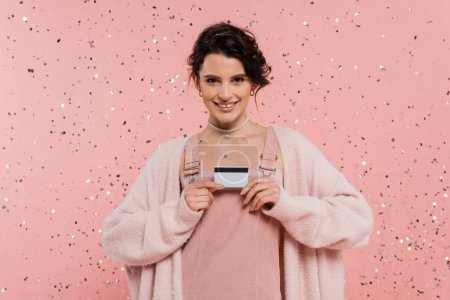 brünette Frau in trendiger Kleidung zeigt Kreditkarte und lächelt in die Kamera unter Konfetti auf rosa Hintergrund
