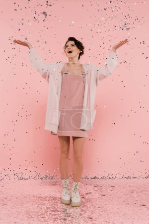 full length of joyful woman in fluffy cardigan catching confetti with raised hands on pink 