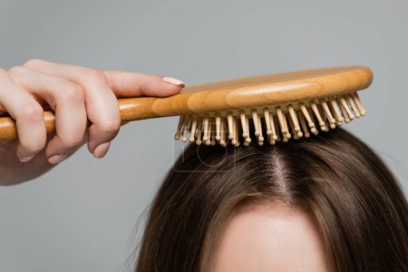 vue recadrée de la jeune femme brossant les cheveux brillants et sains avec brosse à cheveux en bois isolé sur gris