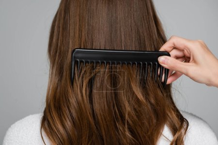 abgeschnittene Ansicht des Friseurs Bürsten glänzende und gesunde Haare der jungen Frau mit Kamm isoliert auf grau