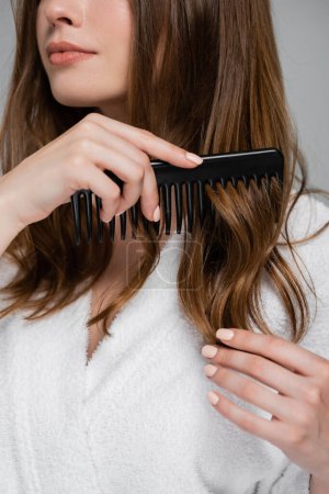 Téléchargez les photos : Vue recadrée de la jeune femme brossant les cheveux brillants et sains avec peigne en plastique isolé sur gris - en image libre de droit