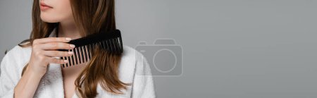 Téléchargez les photos : Vue partielle de la jeune femme brossant les cheveux brillants avec peigne en plastique isolé sur gris, bannière - en image libre de droit