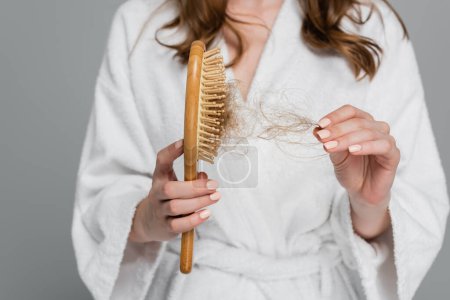 cropped view of young woman holding wooden hair brush and pulling damaged hair isolated on grey 