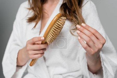 partial view of young woman holding wooden hair brush while pulling damaged hair isolated on grey 