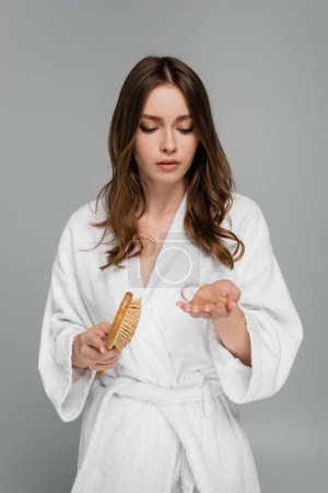 sad young woman in bathrobe holding hair brush and damaged hair isolated on grey 