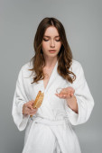 sad young woman in bathrobe holding hair brush and damaged hair isolated on grey  puzzle #627883262