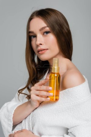 junge Frau mit glänzendem Haar hält Flasche mit Öl isoliert auf grau