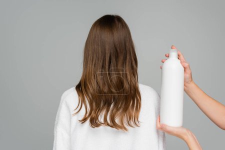 professional hair stylist holding bottle with hair conditioner near young woman isolated on grey