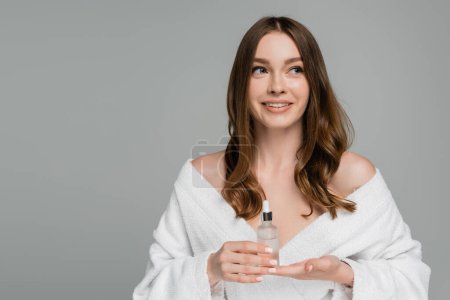 glückliche junge Frau mit glänzendem Haar hält Flasche mit Serum isoliert auf grau 