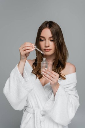 hübsche junge Frau mit glänzendem Haar hält Flasche und Pipette mit Serum isoliert auf grau 