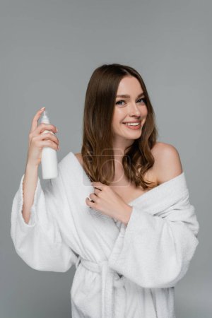 fröhliche junge Frau Styling glänzende Haare mit Haarspray isoliert auf grau 