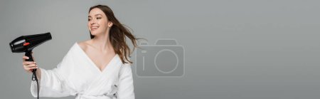 happy young woman with shiny hair using hair dryer isolated on grey, banner 