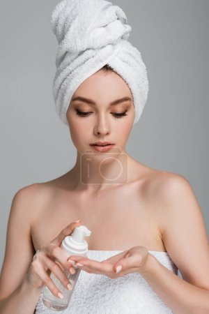 mujer joven con hombros desnudos y toalla en la cabeza sosteniendo la botella con espuma limpiadora aislada en gris 