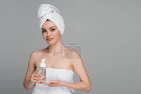 Foto de Joven sonriente con hombros desnudos y toalla en la cabeza sosteniendo la botella con espuma limpiadora aislada en gris - Imagen libre de derechos