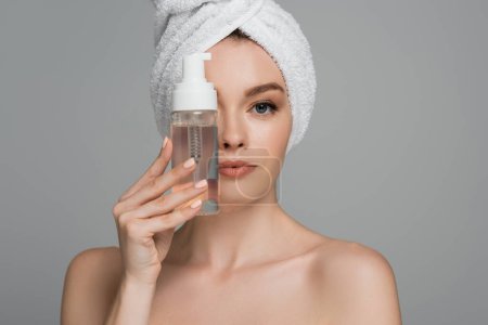 mujer joven con hombros desnudos y toalla en la cabeza sosteniendo botella con espuma limpiadora cerca de la cara aislada en gris 