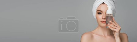 Téléchargez les photos : Jeune femme avec épaules nues et serviette sur la tête tenant bouteille avec mousse nettoyante près du visage isolé sur gris - en image libre de droit