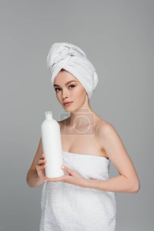 junge Frau mit nackten Schultern und Handtuch auf dem Kopf hält Flasche mit Haarspülung isoliert auf grau 