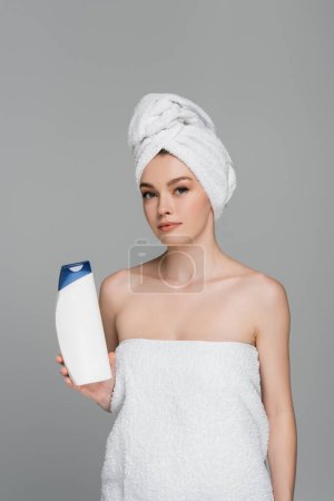 Téléchargez les photos : Jeune femme avec serviette sur la tête tenant bouteille avec shampooing isolé sur gris - en image libre de droit