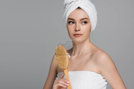 jeune femme avec serviette sur la tête tenant la brosse à cheveux en bois isolé sur gris 