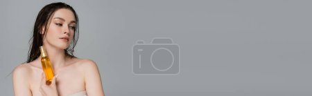 Téléchargez les photos : Jolie jeune femme aux cheveux mouillés et aux épaules nues tenant bouteille avec de l'huile capillaire isolée sur gris, bannière - en image libre de droit