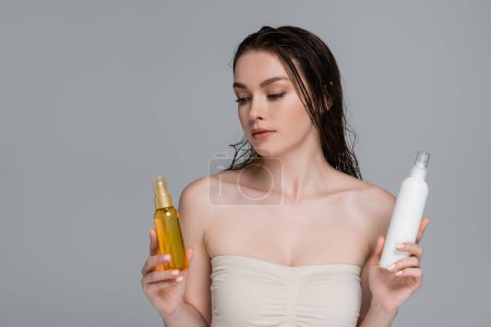 Photo pour Jolie jeune femme aux cheveux mouillés et aux épaules nues au choix entre des flacons de traitement capillaire isolés sur gris - image libre de droit