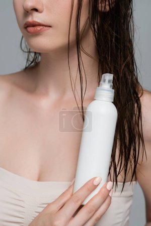 abgeschnittene Ansicht einer jungen Frau mit nackten Schultern, die Sprühflasche mit Behandlung Haarprodukt isoliert auf grau hält 
