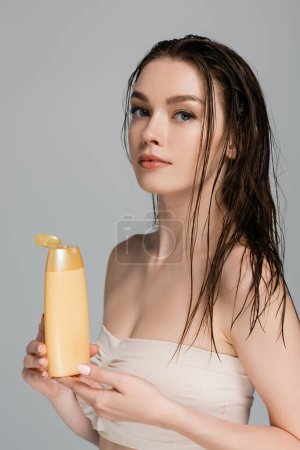Téléchargez les photos : Jolie jeune femme aux cheveux mouillés et épaules nues tenant bouteille avec shampooing isolé sur gris - en image libre de droit