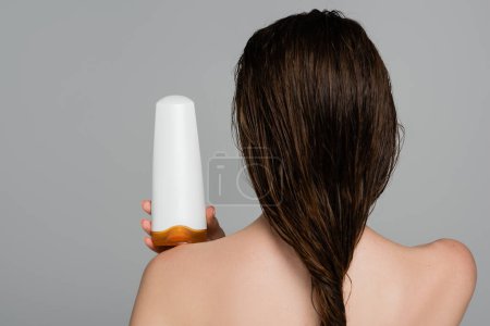 Rückansicht einer jungen Frau mit nassen Haaren und nackten Schultern, die eine Flasche mit Shampoo hält, isoliert auf grau 