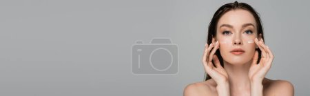 Foto de Mujer joven con el pelo mojado aplicando crema facial en las mejillas aisladas en gris, pancarta - Imagen libre de derechos