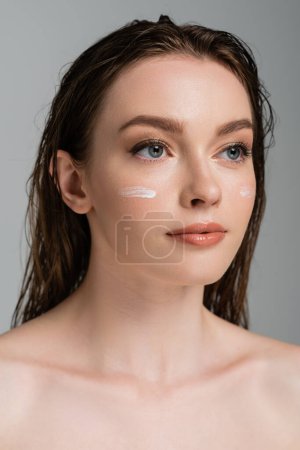 pretty young woman with wet hair and moisturizing cream on cheeks looking away isolated on grey 